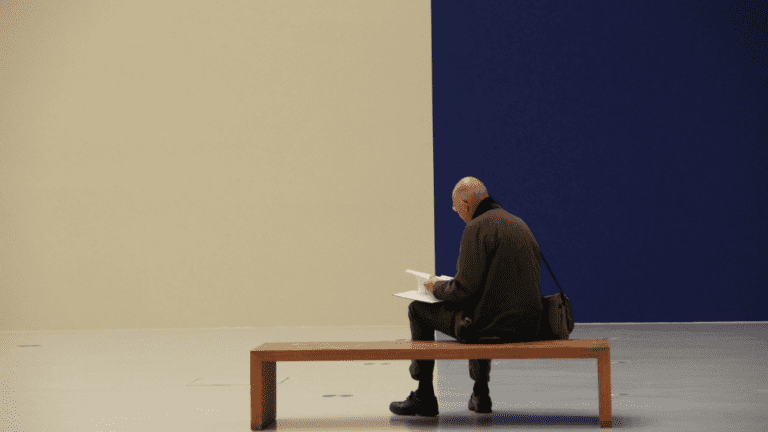 A man sitting on a bench and behind him a wall painted half blue half sepia.