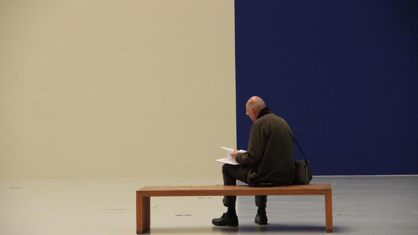 A man sitting on a bench and behind him a wall painted half blue half sepia.